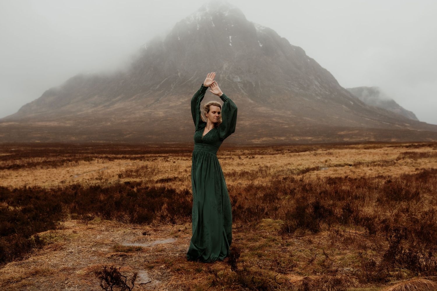 Woman in green dress poses arms raised before misty mountain in Scottish Highlands moody landscape.