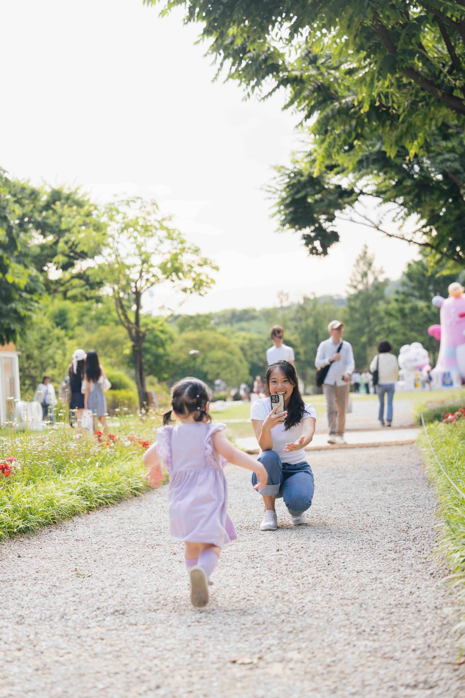 A mother crouches on a park path photographing her young daughter in a purple dress running toward her.