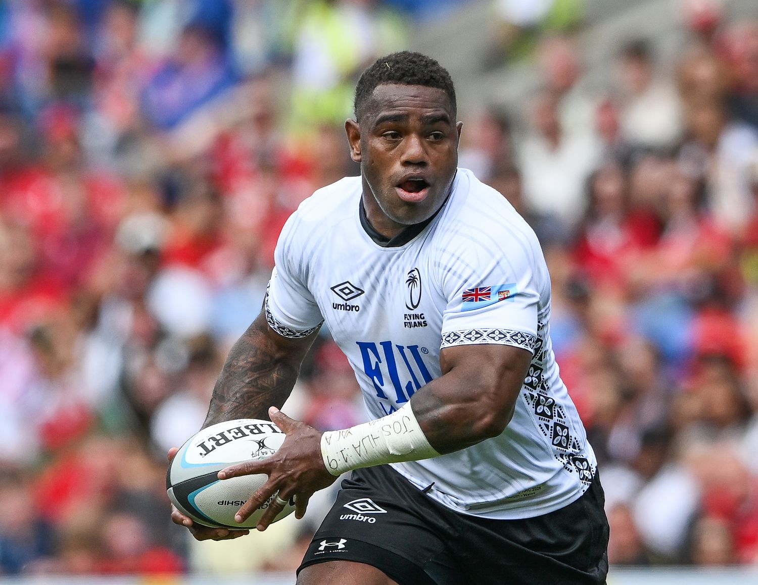 Fijian rugby player in white Umbro jersey runs with ball during a sunny outdoor match.