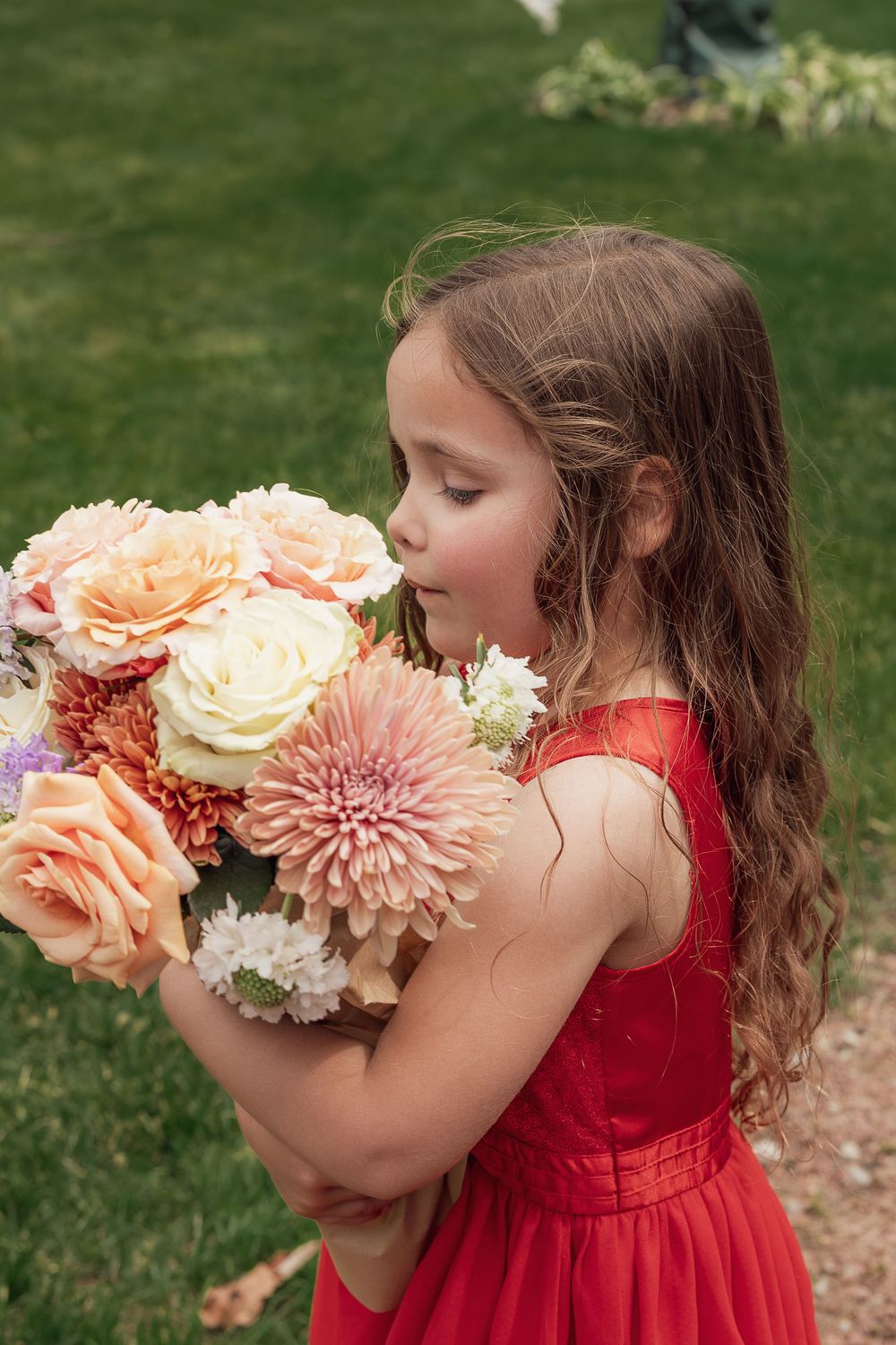 Young girl in red dress smelling a large bouquet of colorful flowers outdoors on green grass.