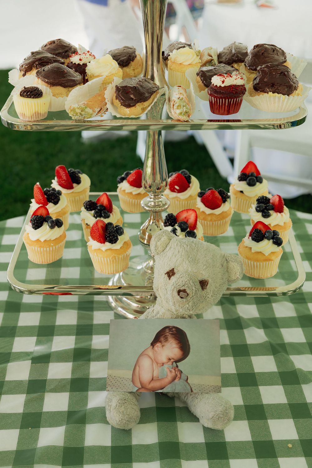 Three-tier dessert stand with berry-topped cupcakes, chocolate treats, and a teddy bear with baby photo at a baby shower.
