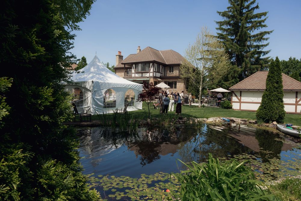 Elegant outdoor event tent beside a serene pond with lush gardens and a large Tudor-style home in the background.