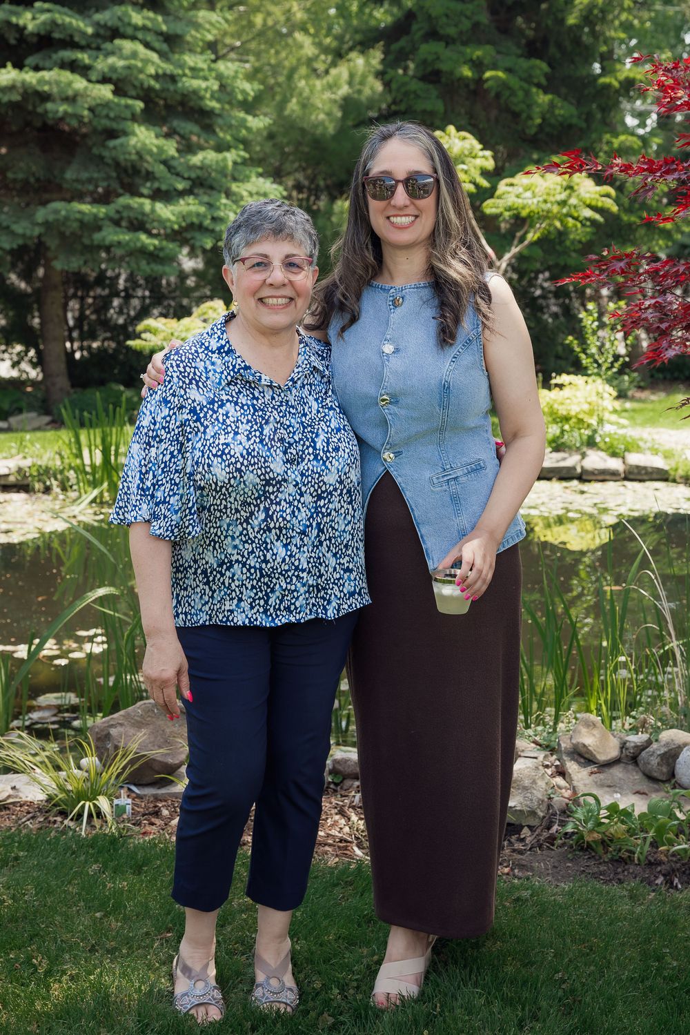 Two women smiling together outdoors in a garden setting, one older in floral top, one younger in denim vest and sunglasses.