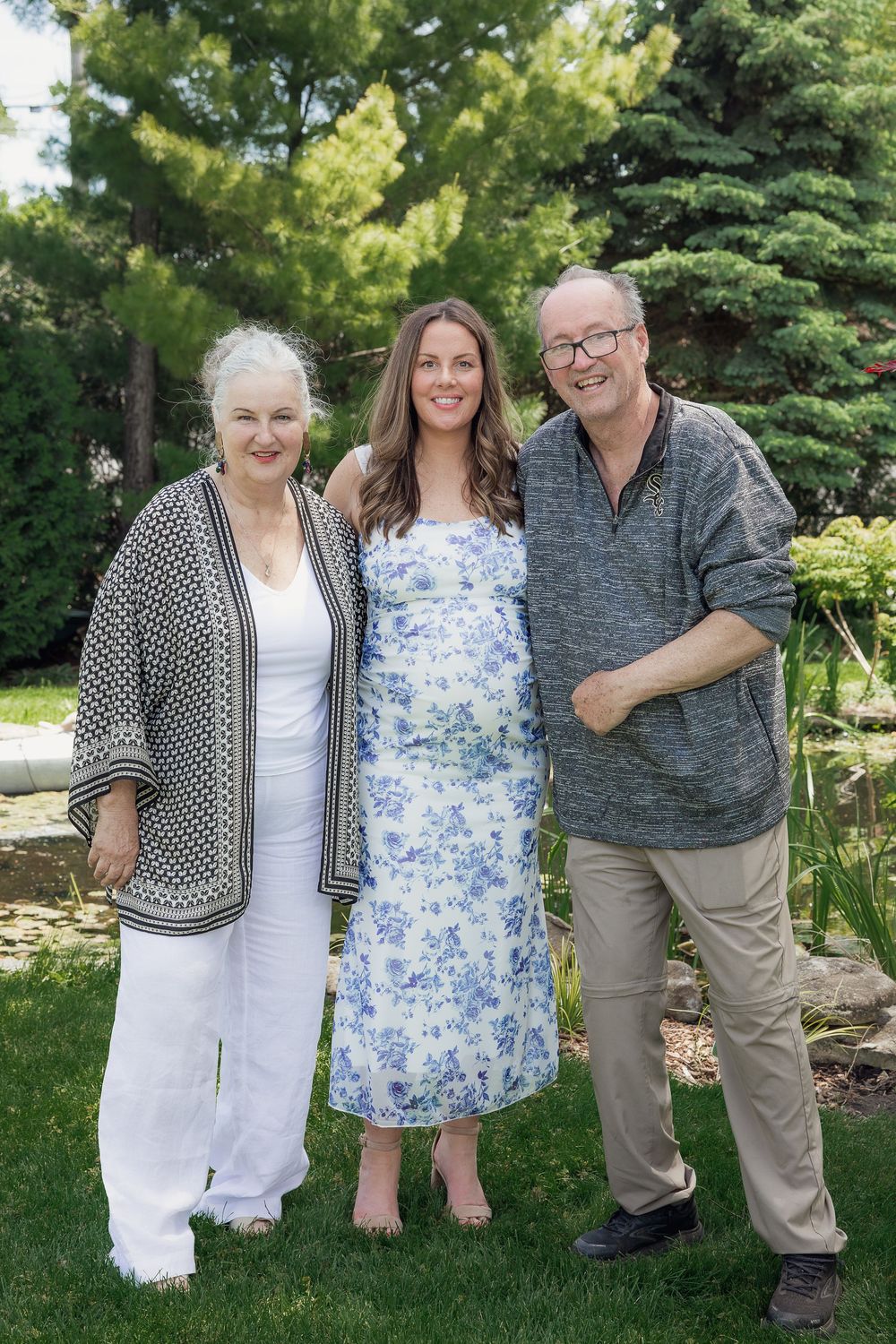 Three people smiling outdoors in a lush garden, two older adults flanking a young woman in a floral dress.