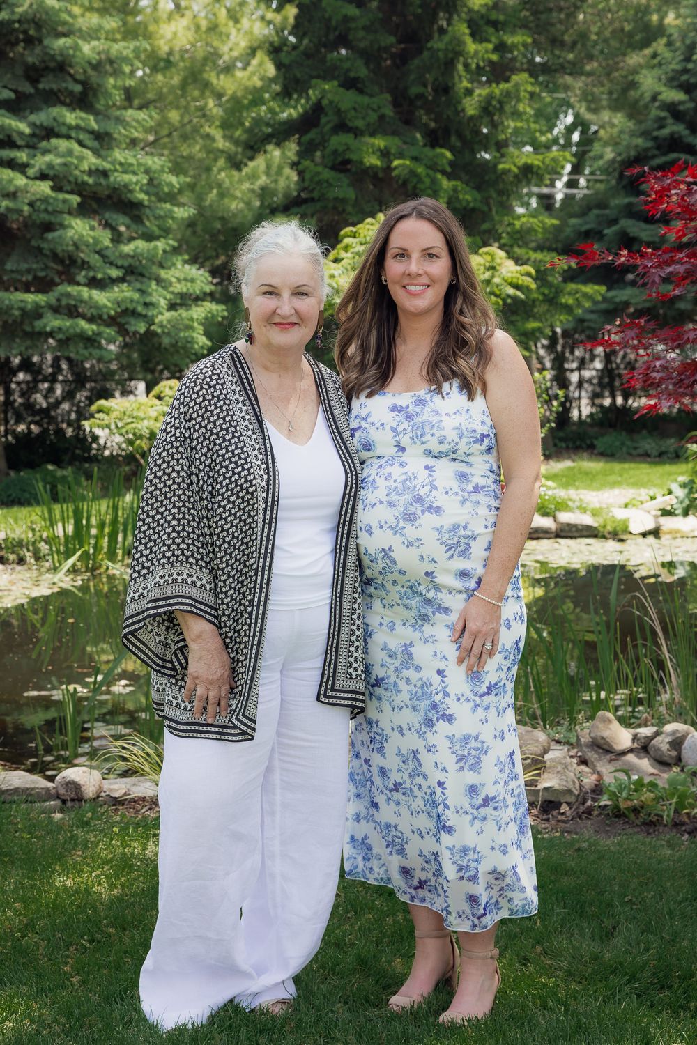 Two women, one elderly and one pregnant, pose together in a lush garden wearing elegant white and floral dresses.