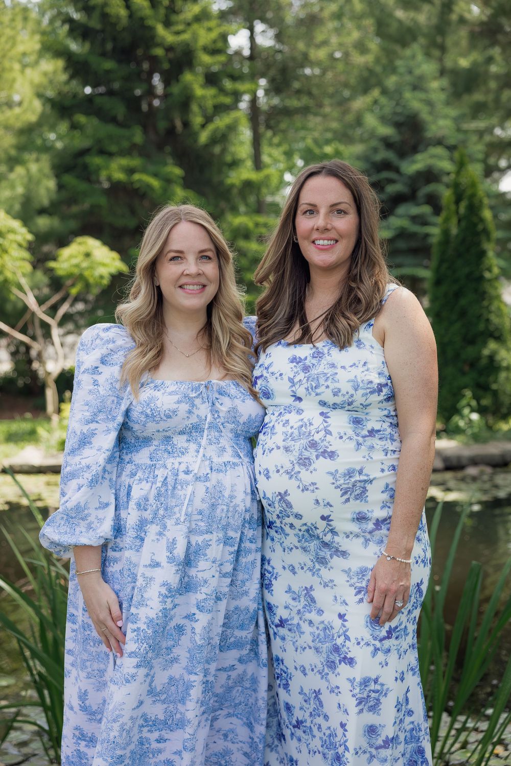 Two pregnant women in matching blue floral dresses smiling together outdoors in a lush garden setting.
