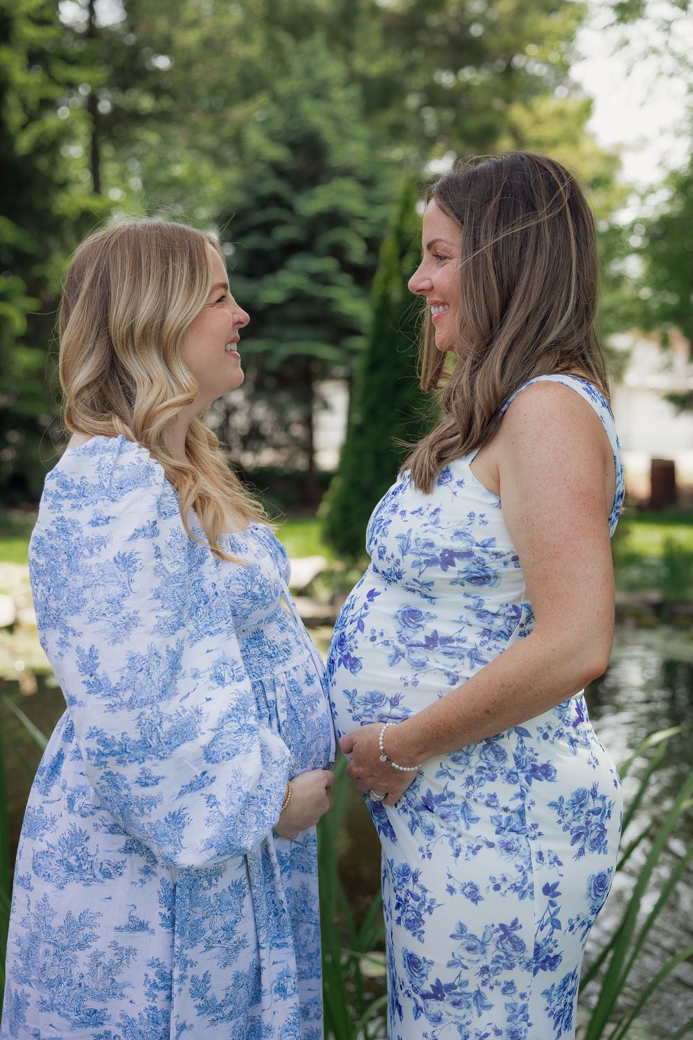Two pregnant women in matching blue floral dresses smiling at each other outdoors near a pond.