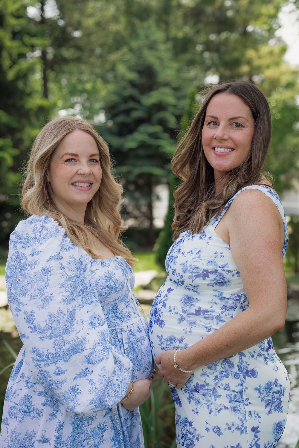 Two smiling pregnant women in blue floral dresses posing outdoors in a green garden setting.