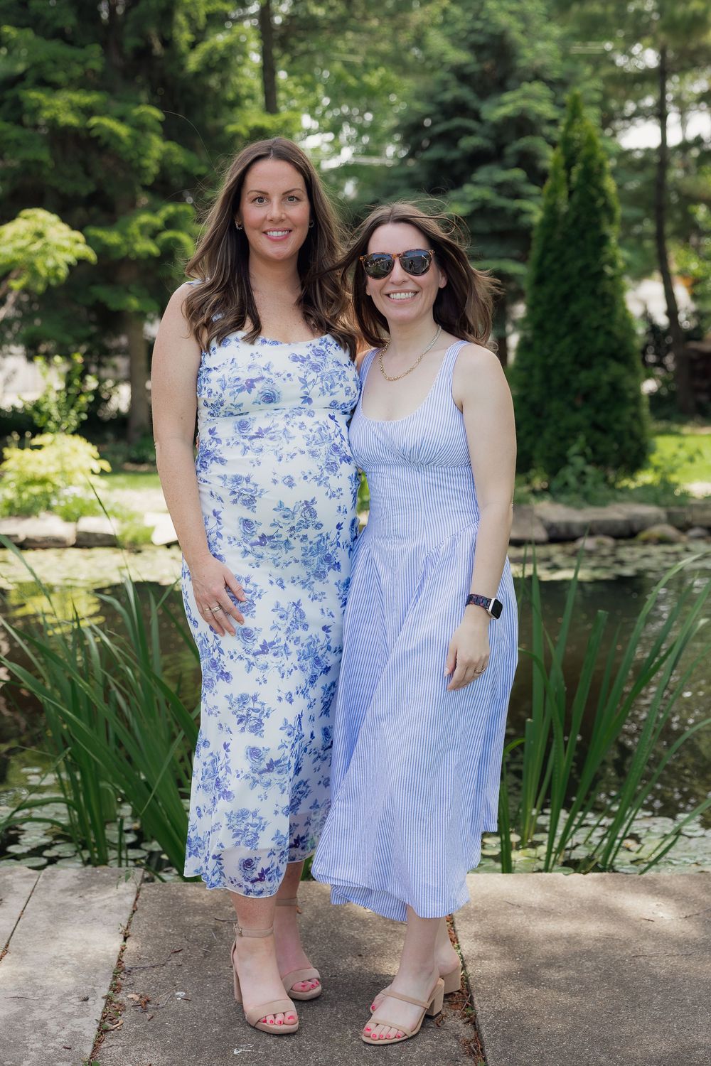 Two women in blue dresses pose together in a lush garden near a pond on a sunny day.