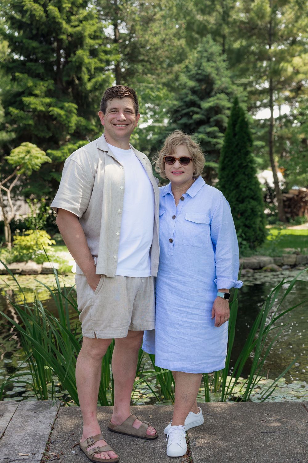 A smiling couple poses outdoors near a pond surrounded by lush green trees on a sunny day.