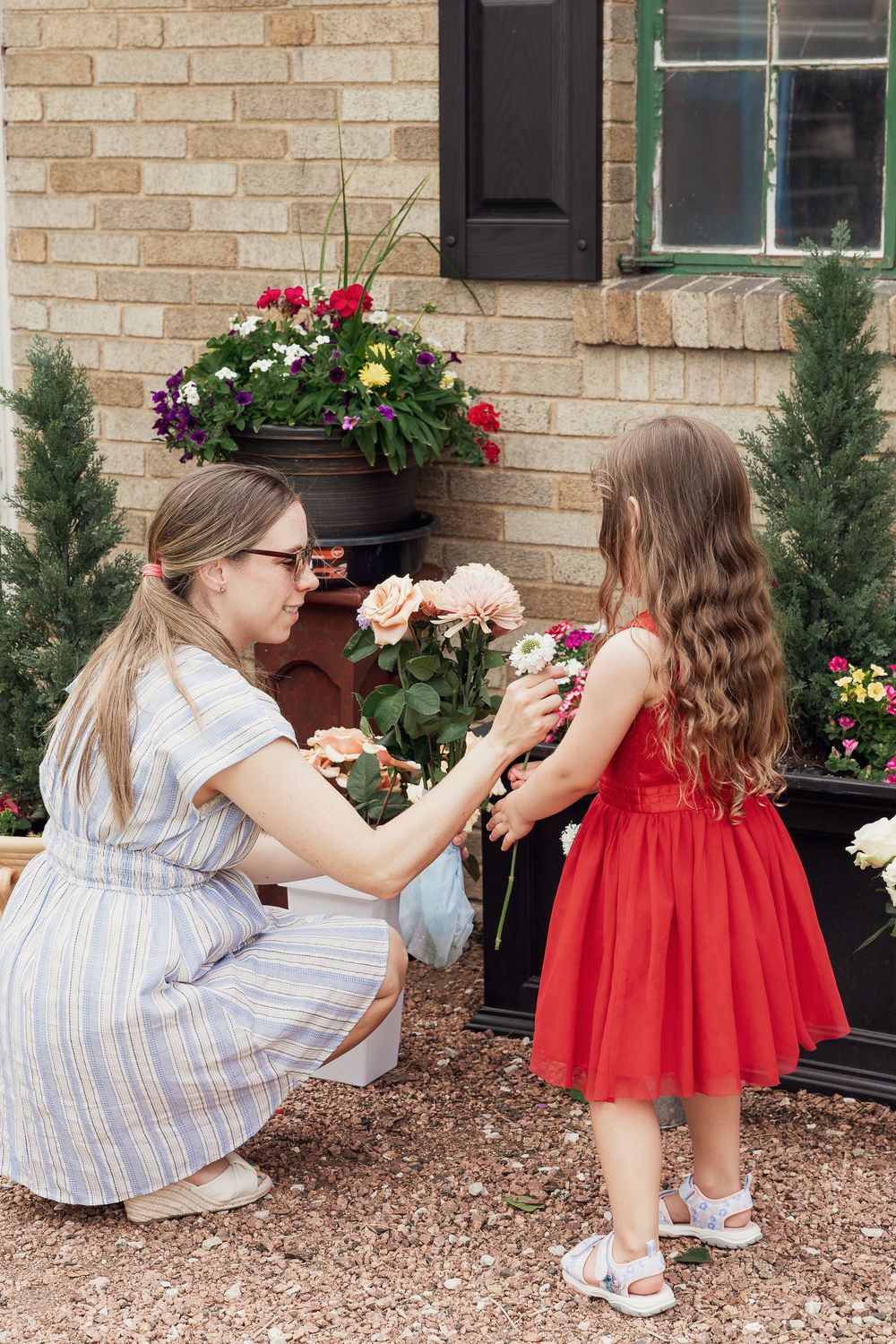 A woman in a blue dress kneels to show flowers to a little girl in a red dress outside a brick home.