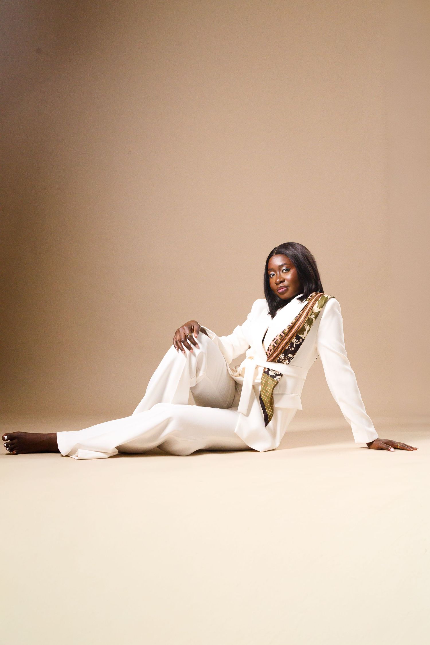 A Black woman in a white pantsuit with a gold patterned sash sits gracefully on a beige studio floor.