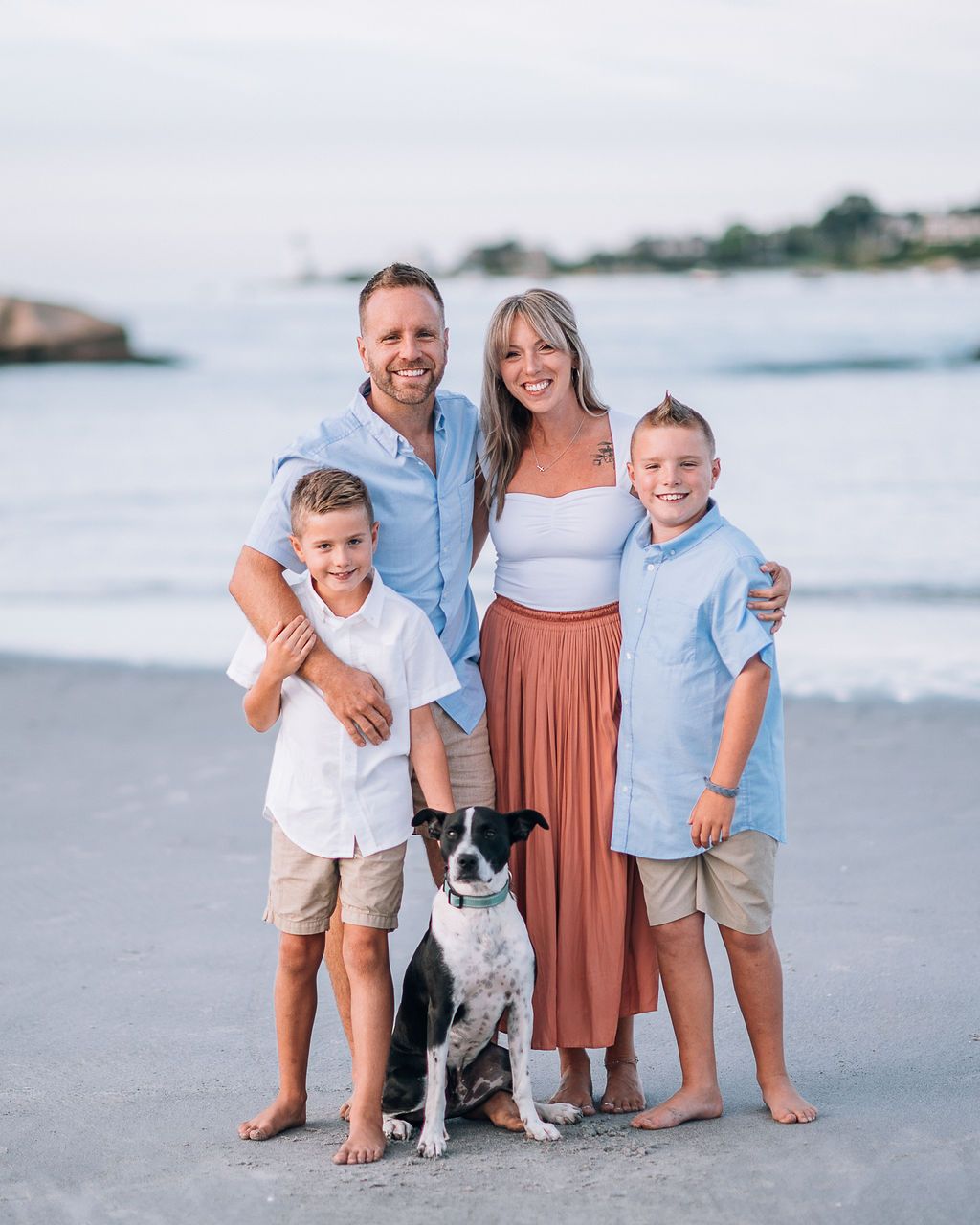 Happy family of four with a dog posing on a beach at sunset, wearing casual summer outfits.