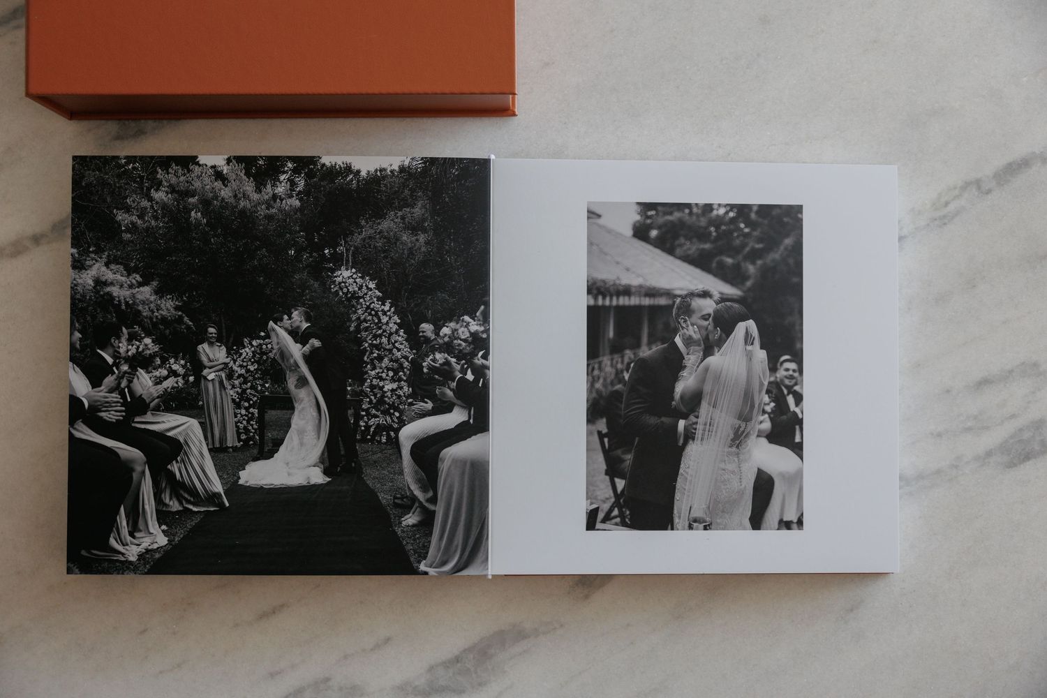 Open wedding photo album on marble surface showing black and white ceremony images, with orange hardcover box beside it.