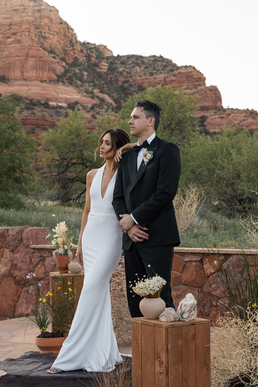Couple in wedding attire poses against Sedona red rock backdrop, bride in white gown, groom in black tuxedo.