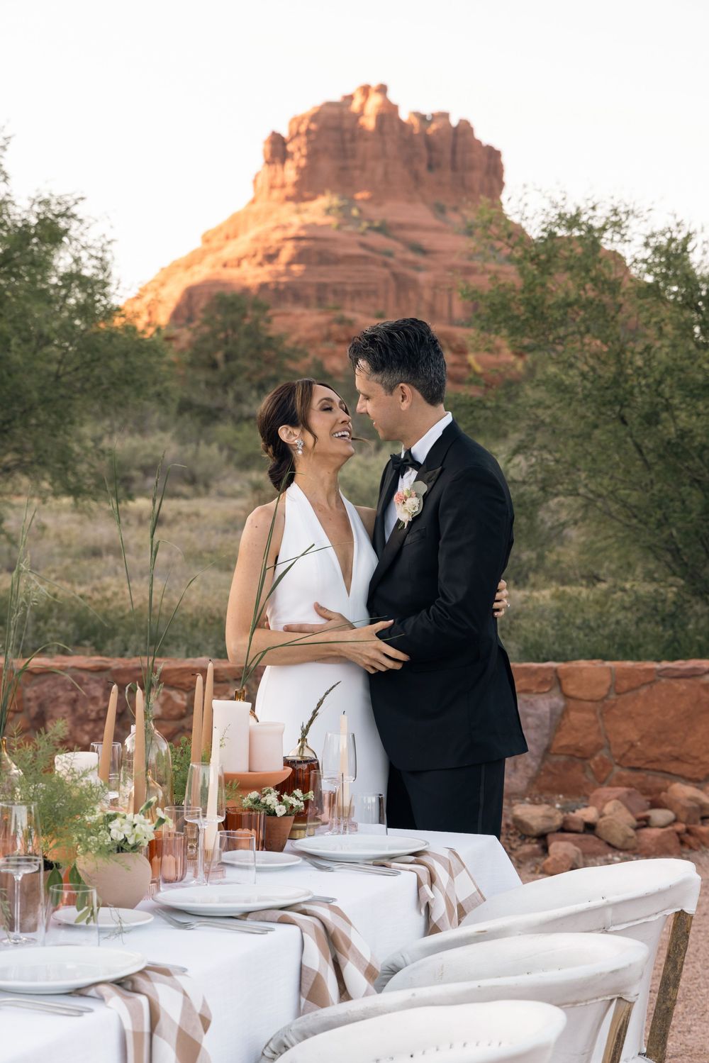 A couple in wedding attire embraces near a decorated table with Bell Rock red formation backdrop in Sedona, Arizona.