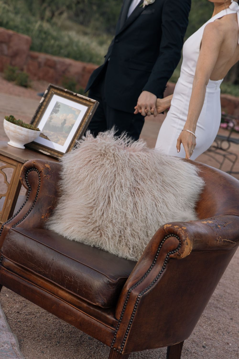 Bride and groom holding hands at outdoor wedding ceremony beside vintage leather armchair with fluffy fur pillow.