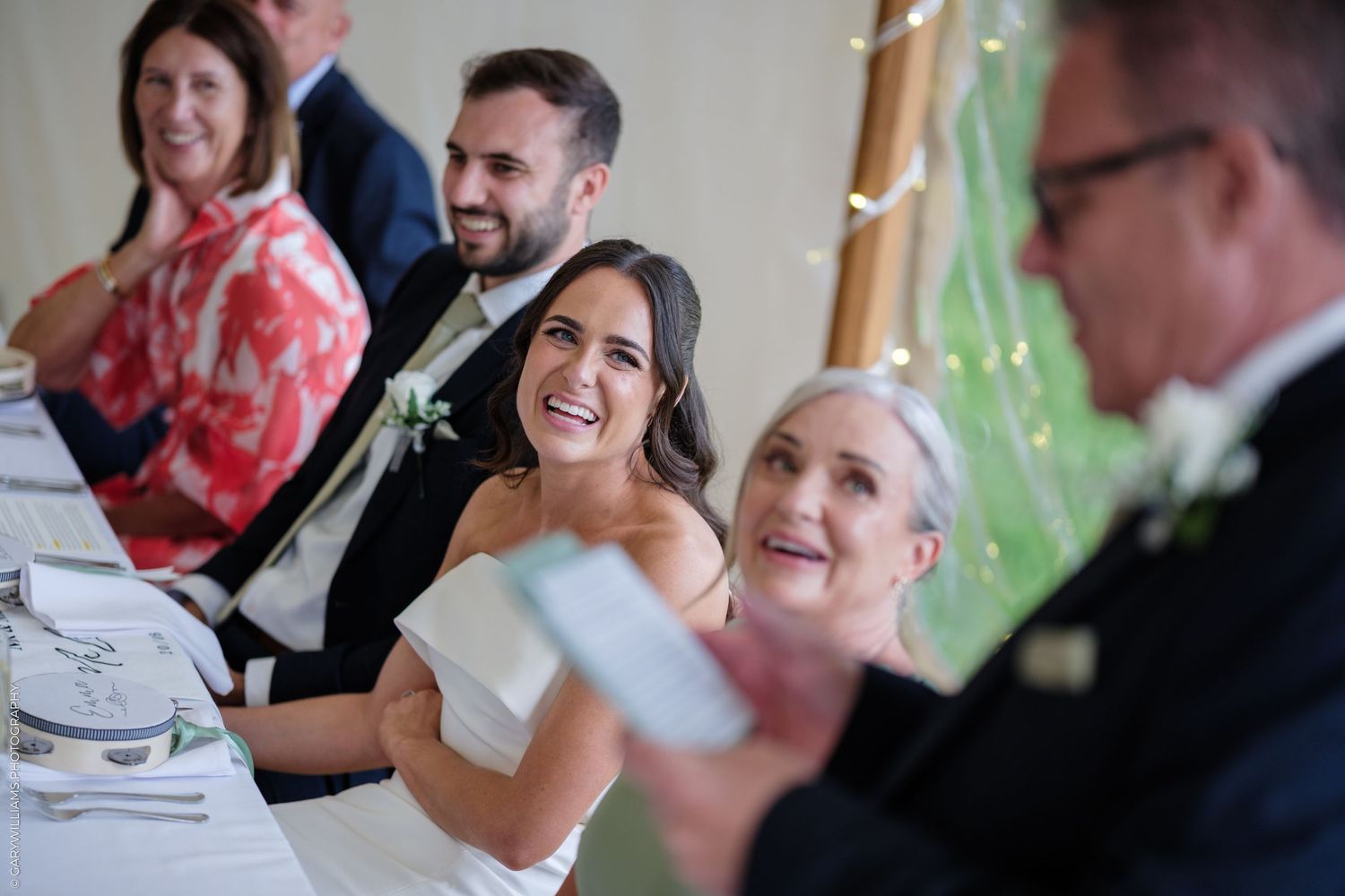 Bride laughing joyfully at wedding reception table during speeches, with guests smiling in background.