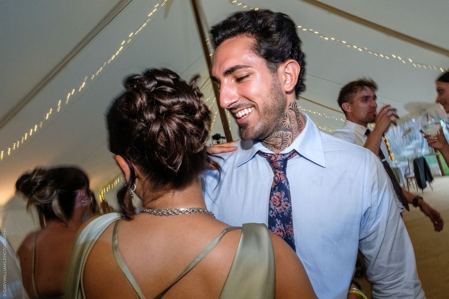 Couple dancing closely at a wedding reception under a tent with string lights, man wearing a floral tie and blue shirt.