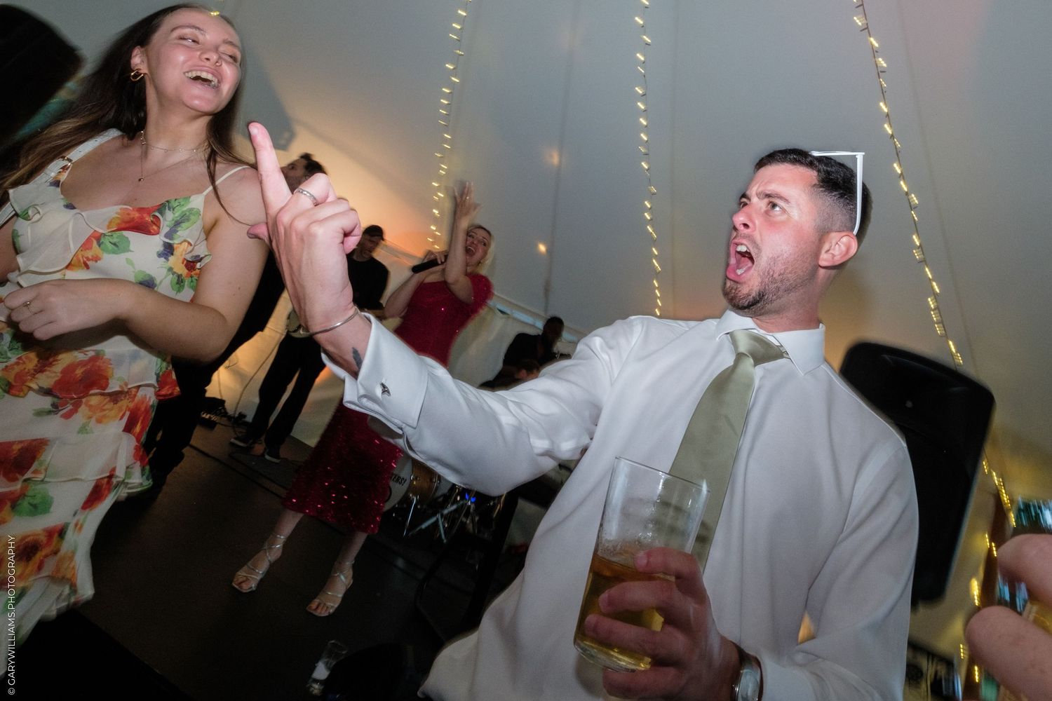 Couple dancing energetically at a wedding reception, man holding drink, woman in floral dress, string lights in background.