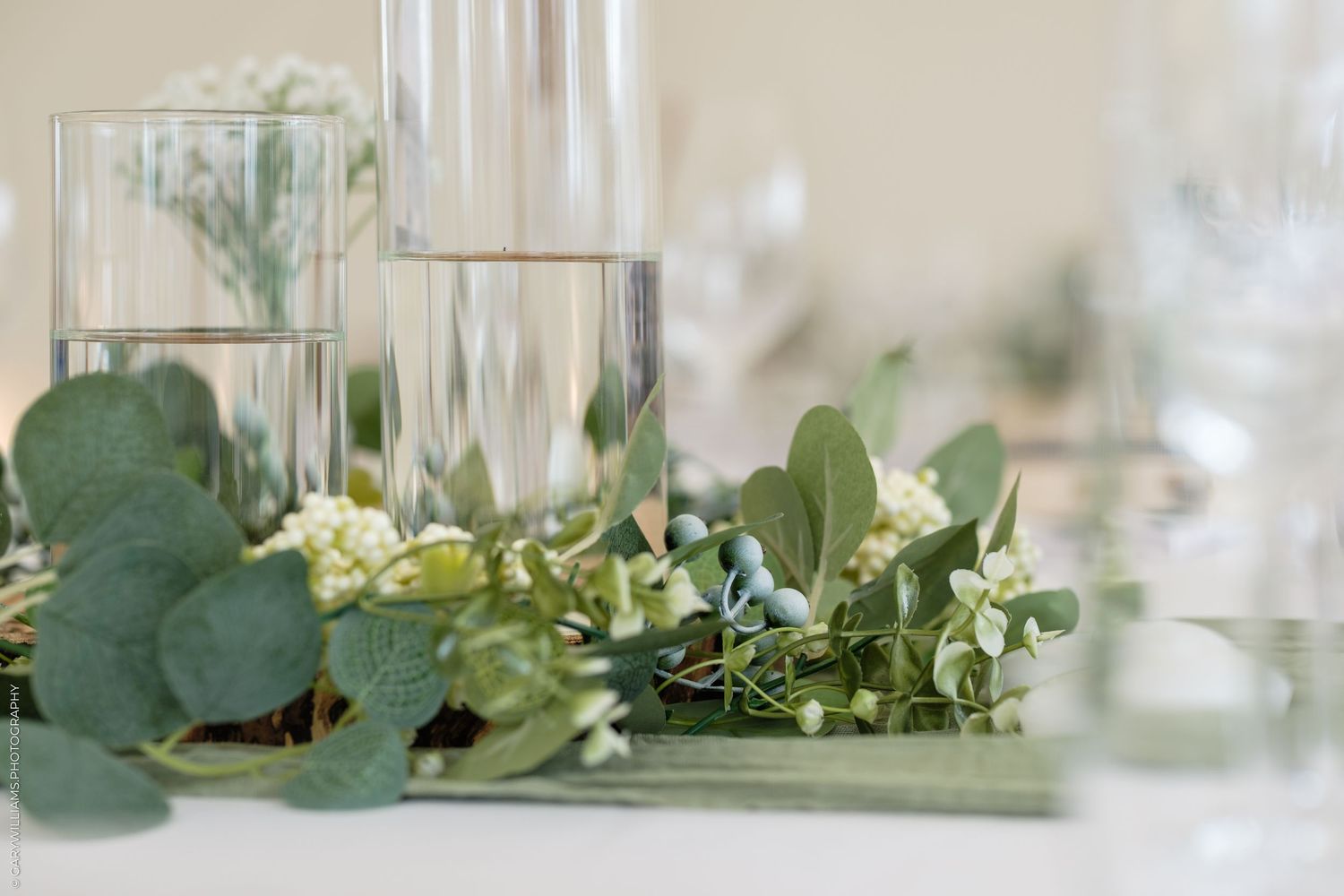 Elegant wedding table centerpiece with glass vases, eucalyptus, and white flowers on a white tablecloth.