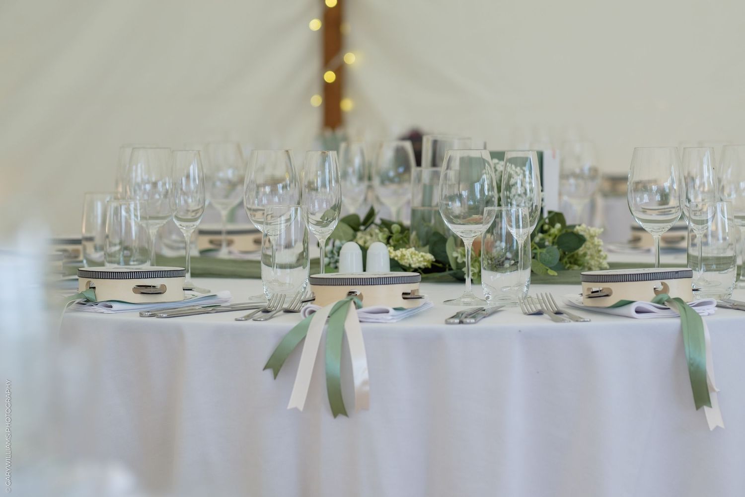 Elegant wedding reception table set with crystal glasses, white linens, and sage green ribbon accents in a marquee.