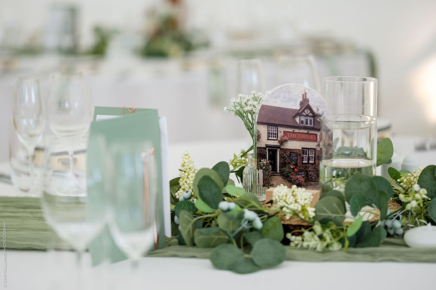 Elegant wedding table centerpiece with miniature house, eucalyptus greenery, and crystal glassware on sage green runner.