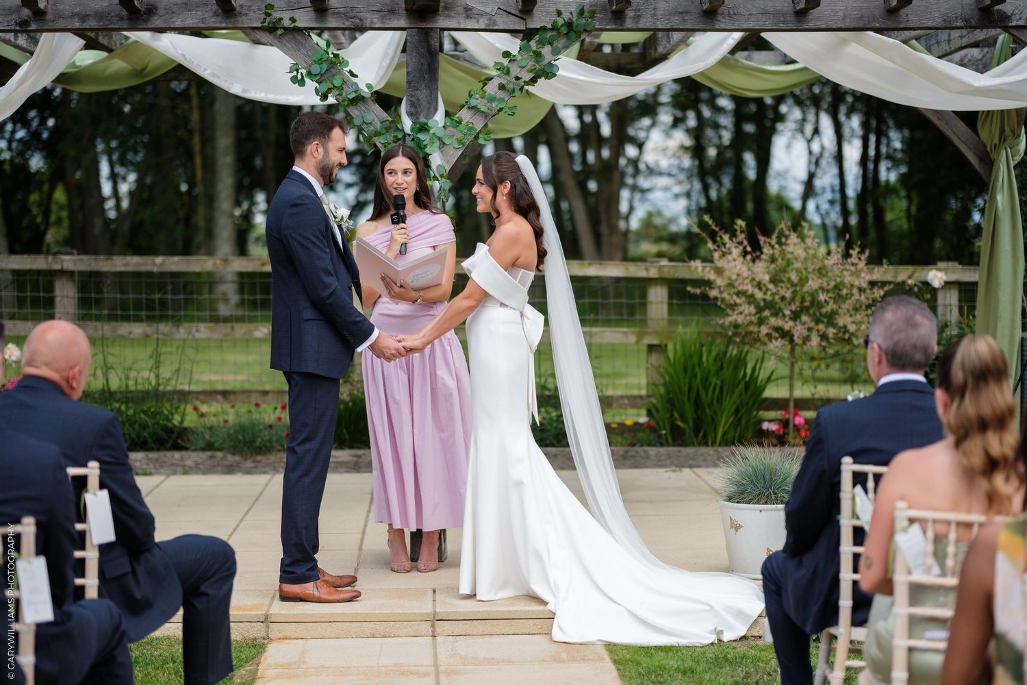 Couple exchanging vows at outdoor wedding ceremony under floral arch with officiant in pink dress.