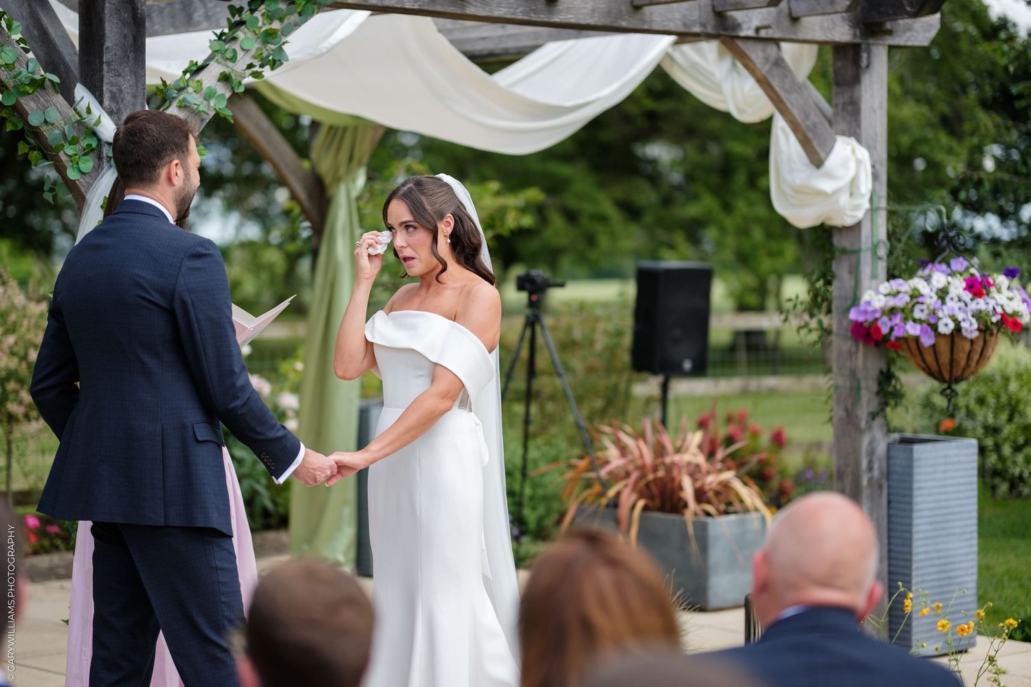 Bride wiping tears during outdoor wedding ceremony under rustic wooden arbor with white draping.