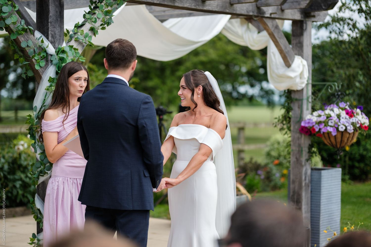 Bride and groom exchange vows under a floral arch with a bridesmaid beside them at an outdoor wedding ceremony.