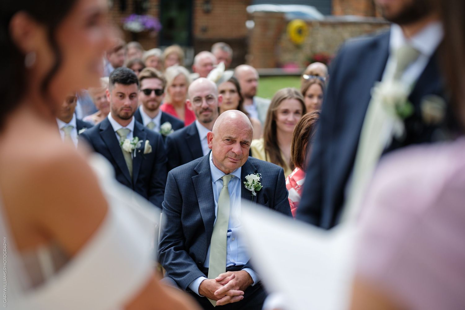 Emotional father watches wedding ceremony outdoors, guests seated behind, wearing navy suit with white buttonhole flower.
