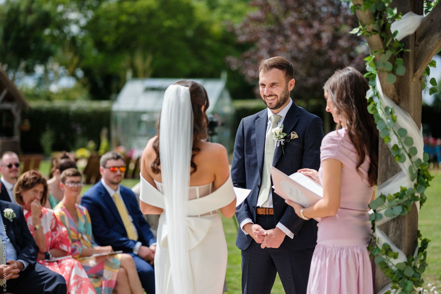Groom smiling at bride during outdoor wedding ceremony with officiant and guests seated in garden.