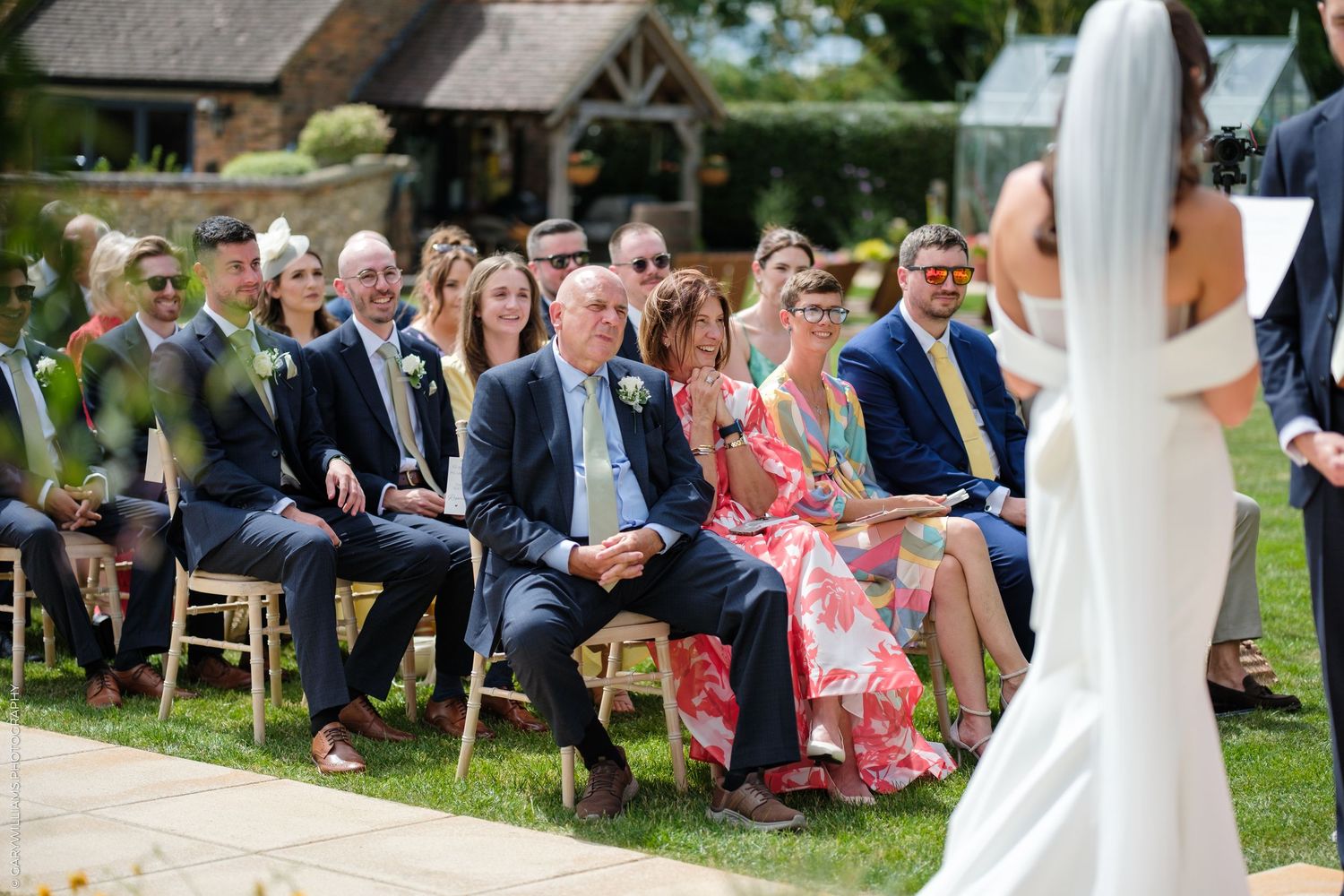 Guests seated outdoors watching a bride walk down the aisle at a sunny garden wedding ceremony.