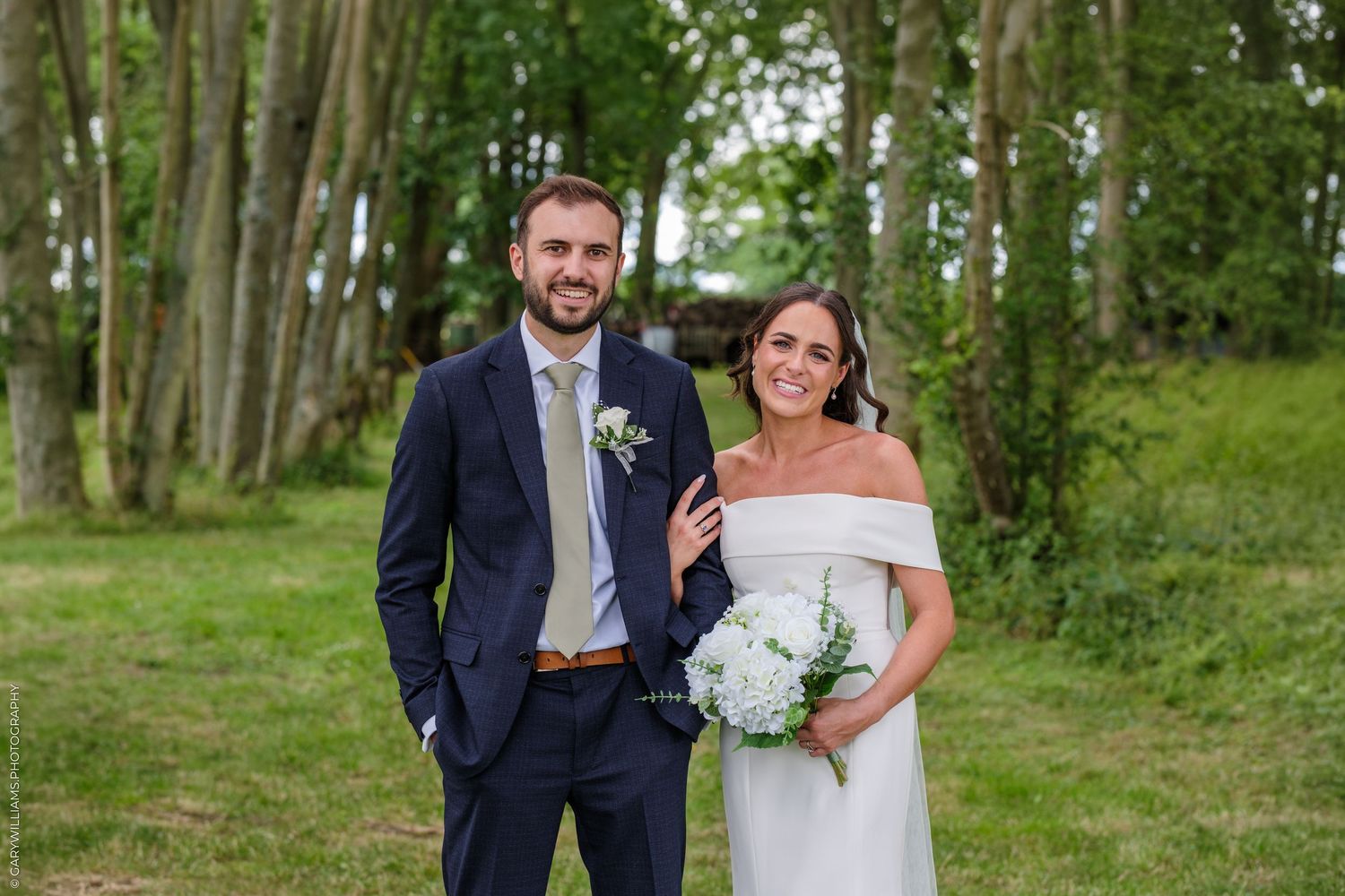 Bride and groom posing outdoors on wedding day, bride holding white bouquet, groom in navy suit, green trees background.