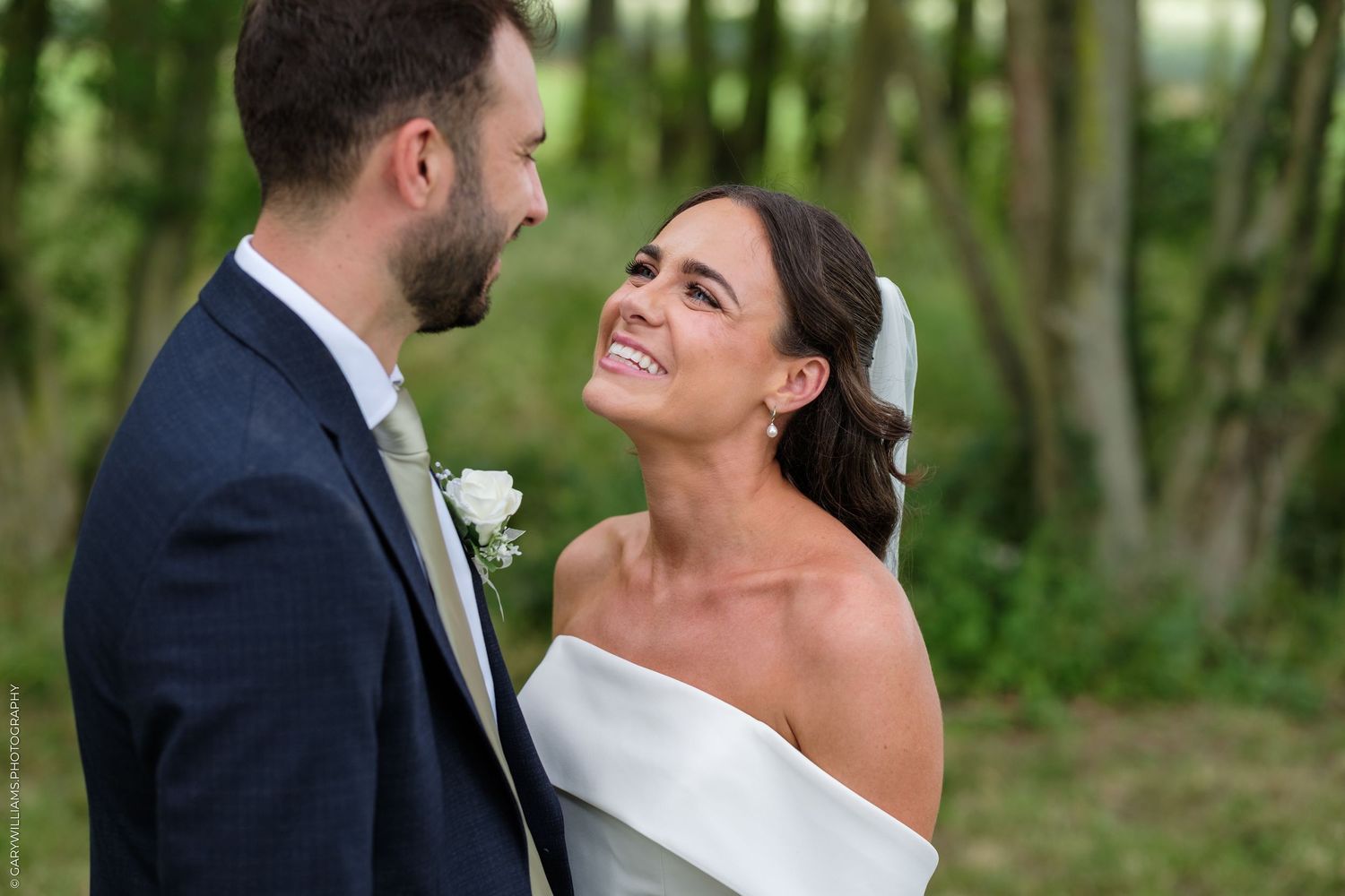 A joyful bride in an off-shoulder white gown smiles at her groom in a navy suit during an outdoor wedding.