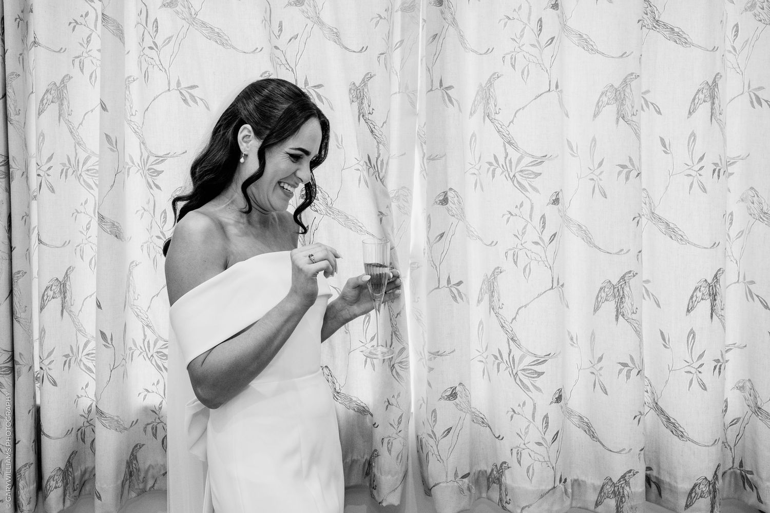 Black and white photo of a smiling bride in a strapless gown reading a note, standing by floral curtains.