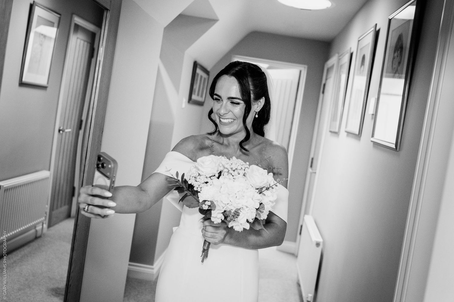 Bride in white gown takes selfie holding bouquet of white flowers in hallway, black and white wedding photo.