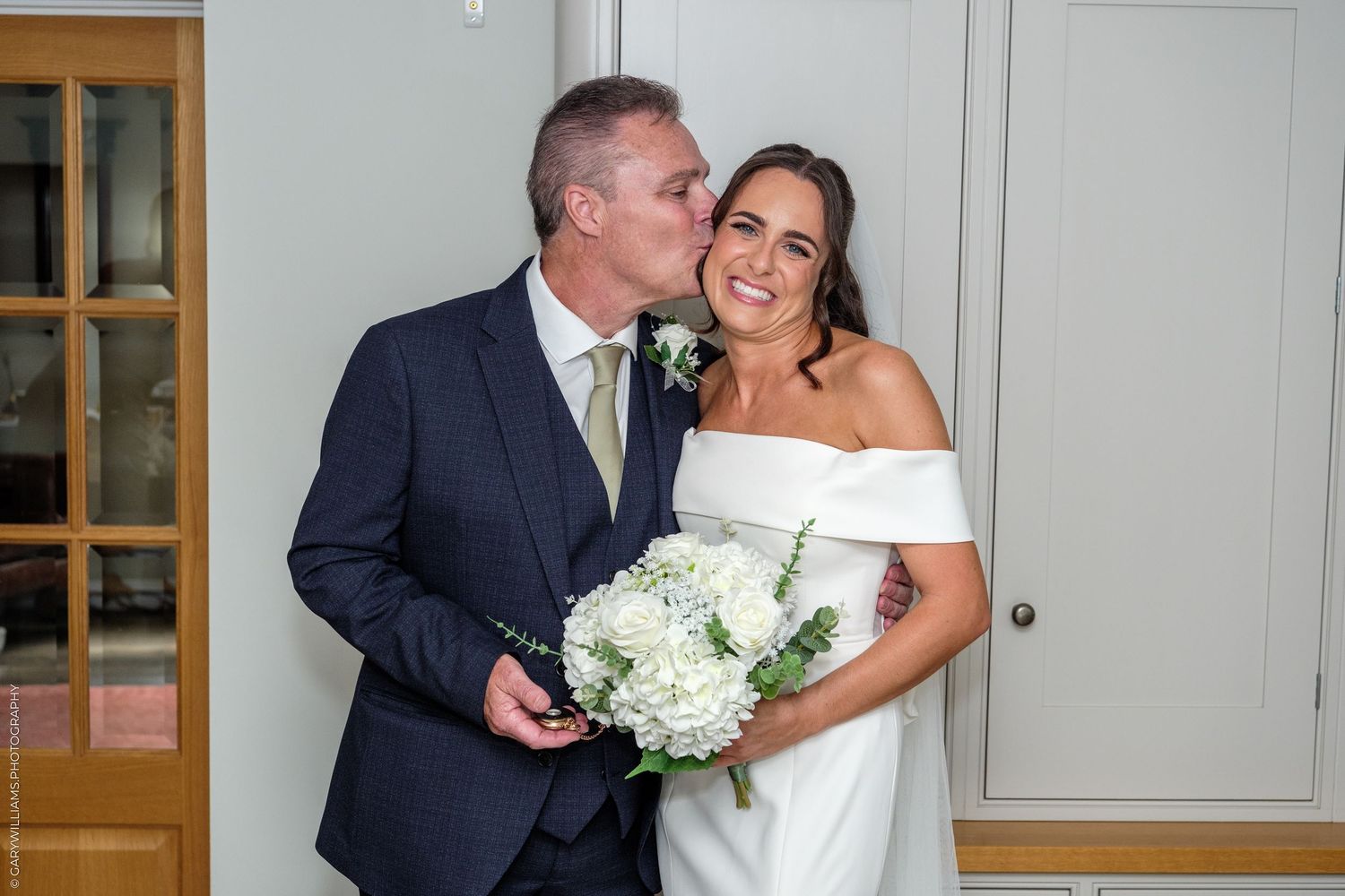 A groom kisses the cheek of his smiling bride holding a white hydrangea bouquet on their wedding day.