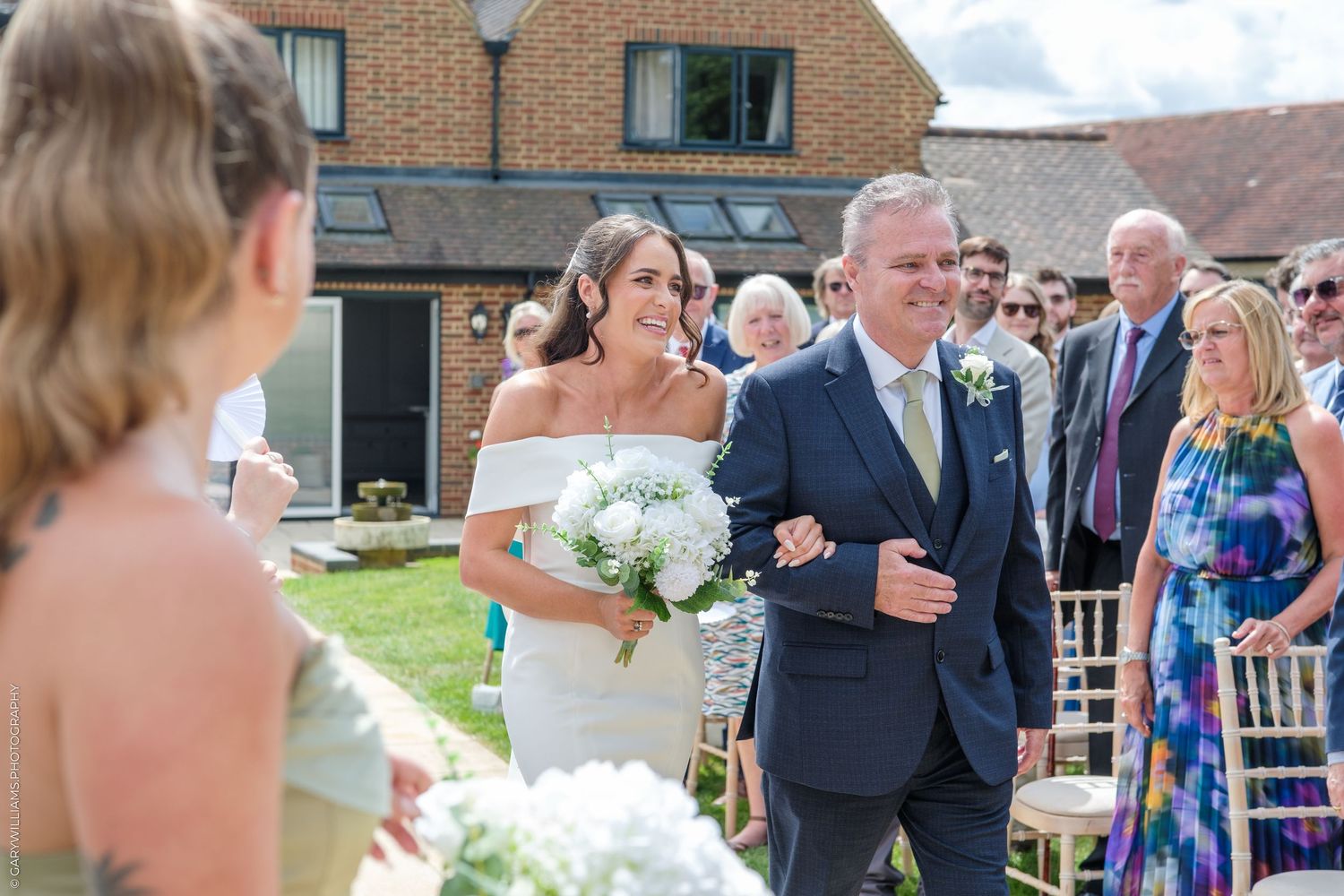 Bride in off-shoulder gown walks down outdoor aisle with father, holding white bouquet at sunny wedding ceremony.