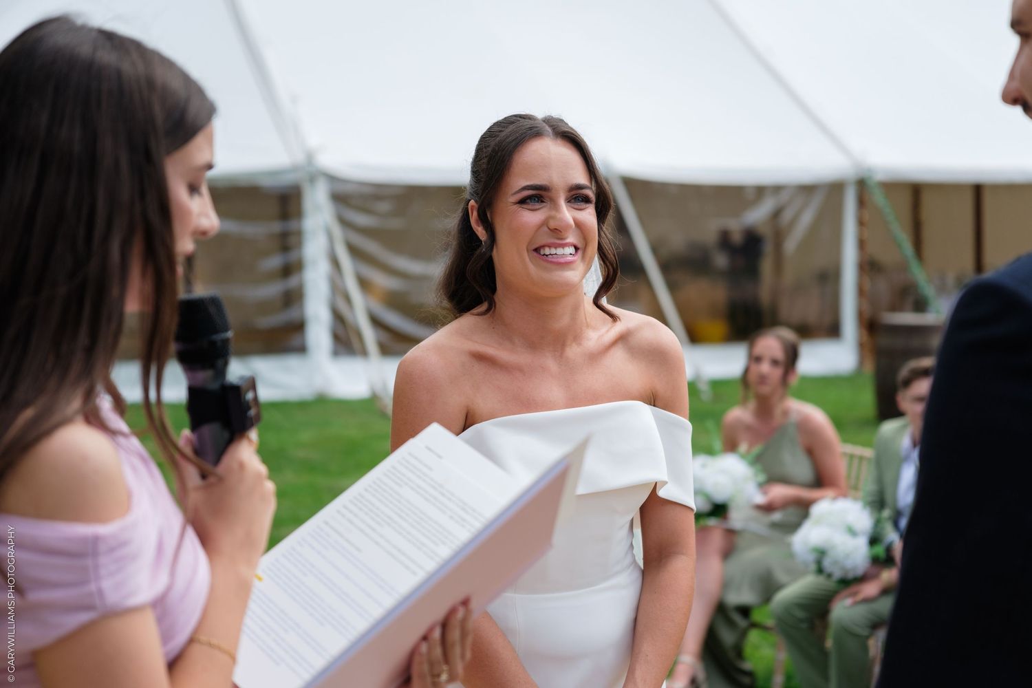 Smiling bride in white off-shoulder gown listening during outdoor wedding ceremony under a white tent.