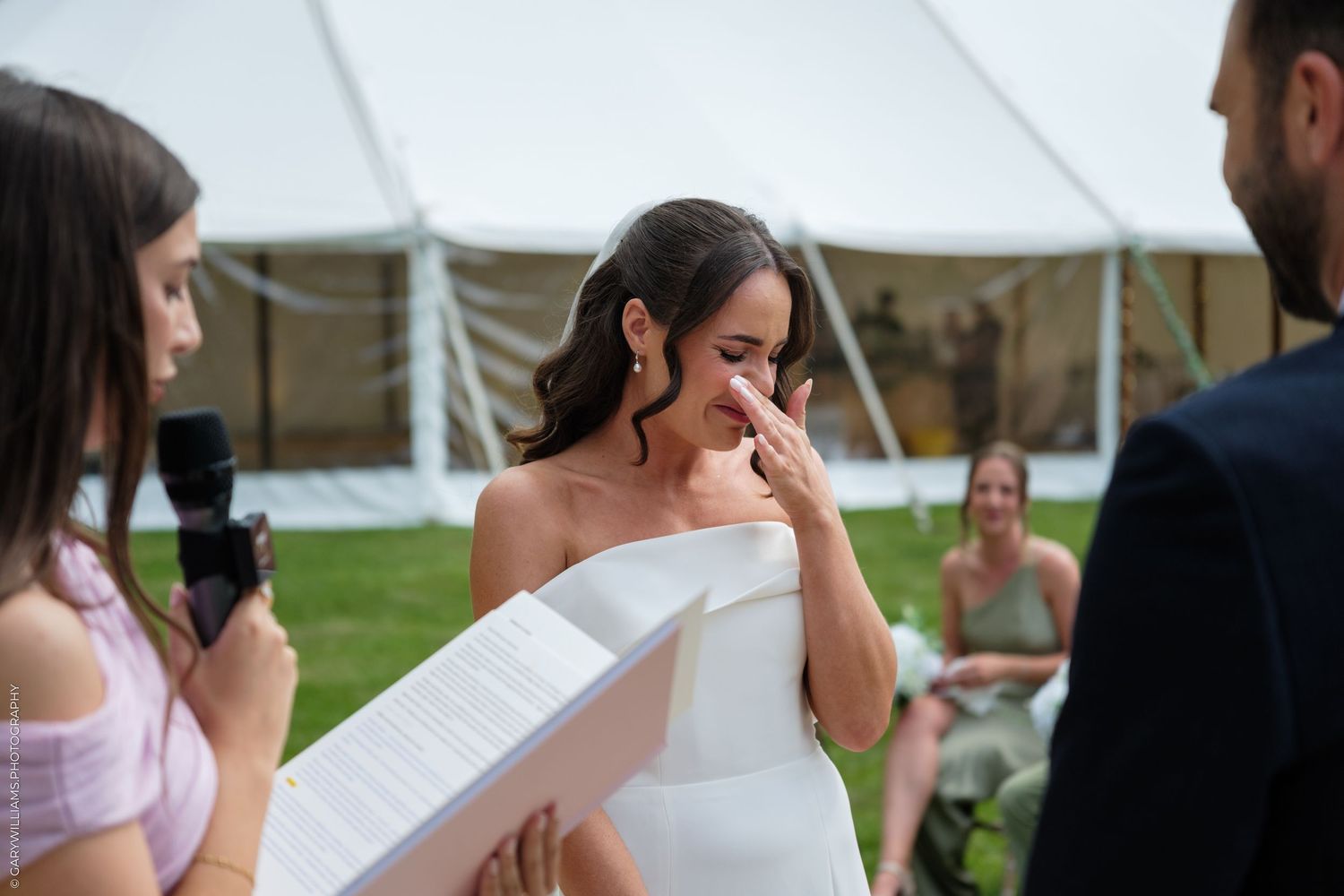 Tearful bride in white dress wiping tears during outdoor wedding ceremony under white tent.