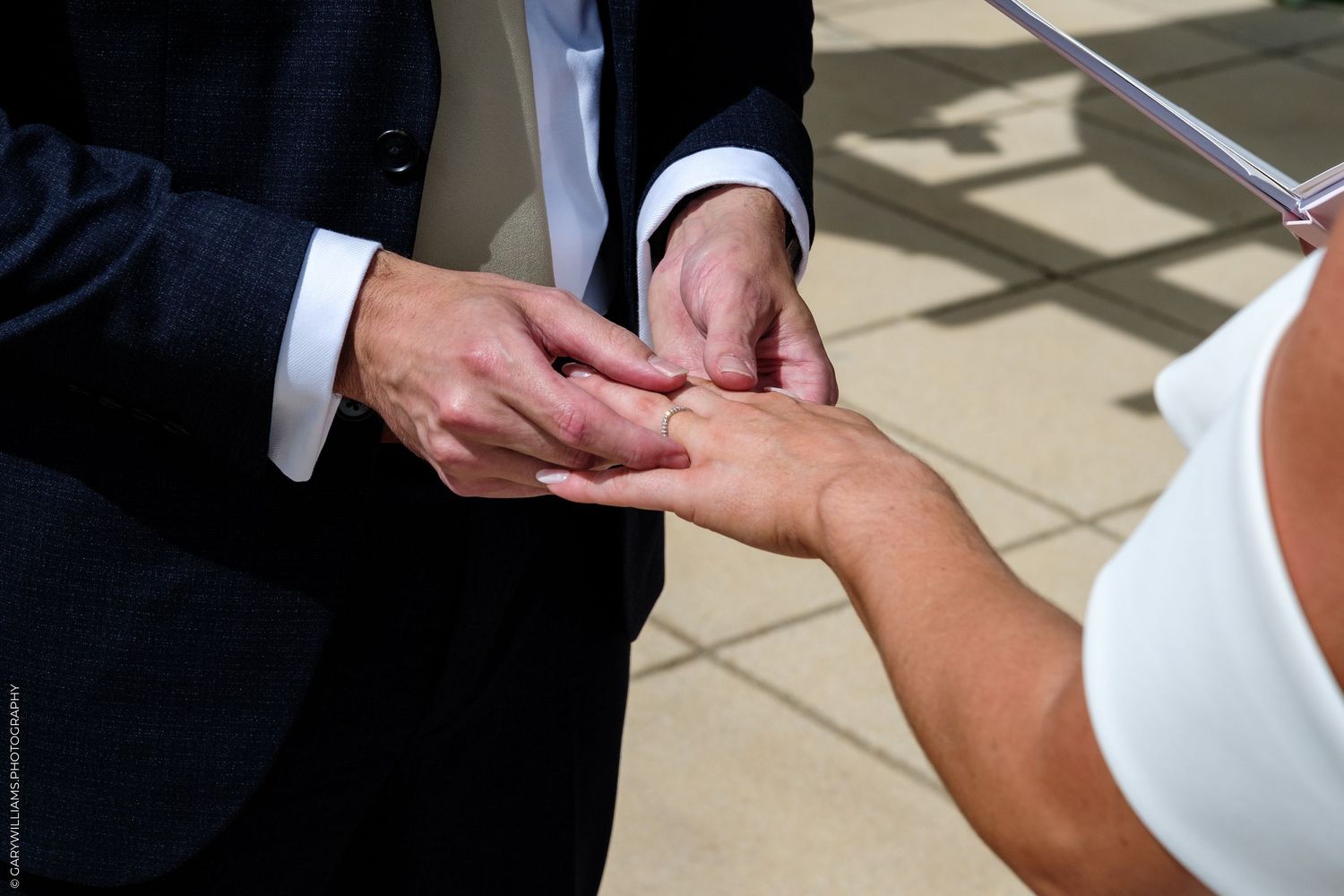 Groom placing a wedding ring on bride's finger during an outdoor ceremony in bright sunlight.