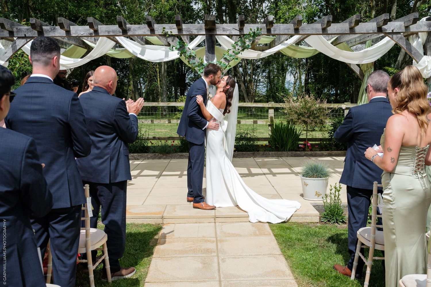 Bride and groom share first kiss at outdoor wedding ceremony with guests and floral pergola backdrop.