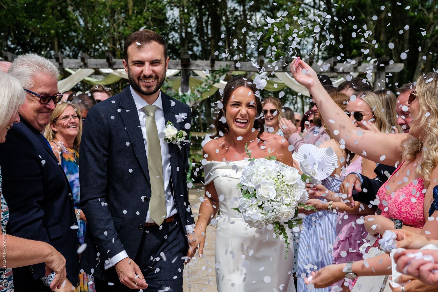 Bride and groom walk through confetti shower surrounded by smiling wedding guests outdoors on a sunny day.