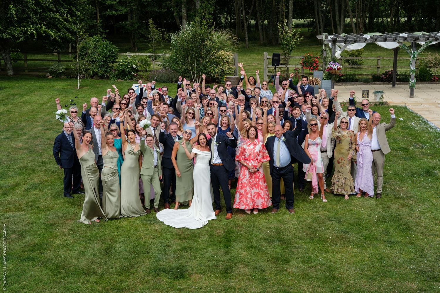 Large wedding group photo outdoors on green lawn, bride in white gown surrounded by elegantly dressed guests celebrating.