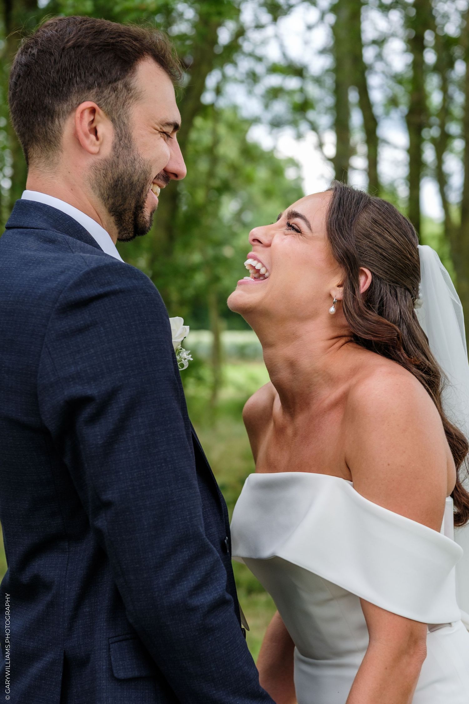 Bride laughing joyfully with groom in blue suit during outdoor wedding portrait among lush green trees.