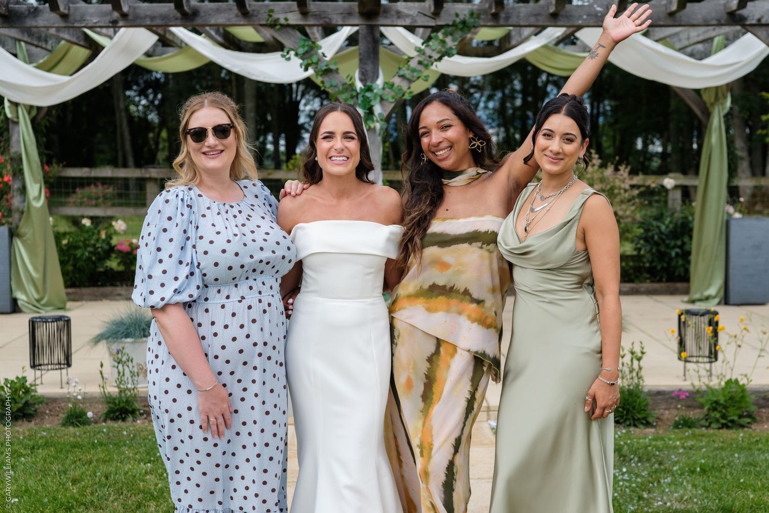 Bride in white gown poses with three bridesmaids in garden wedding venue surrounded by greenery.