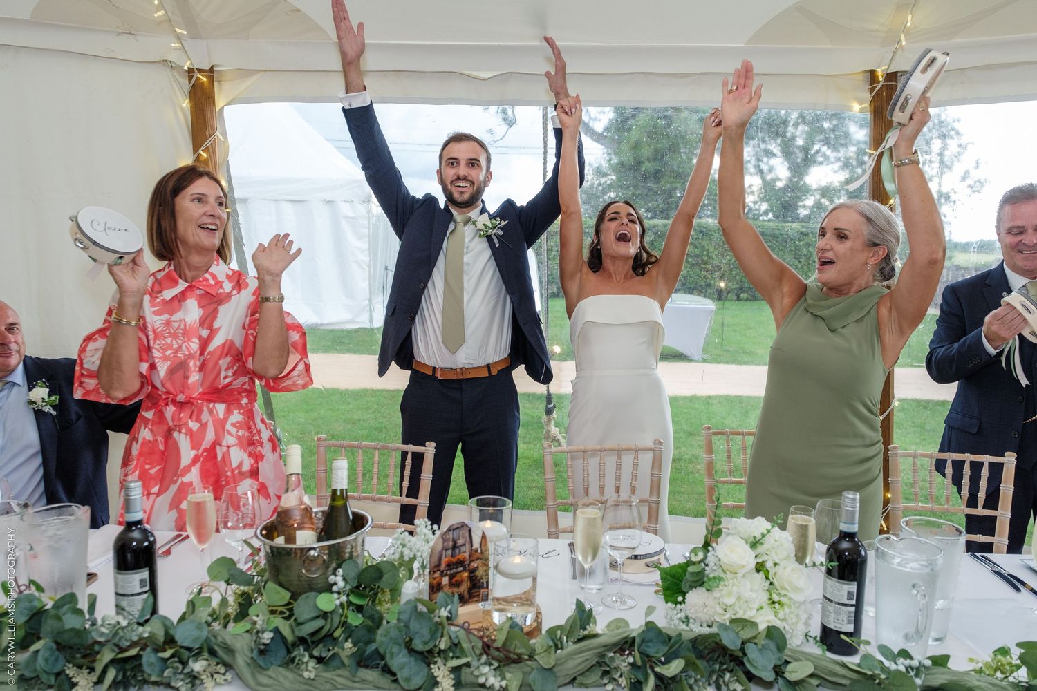 Wedding party celebrating at reception table under marquee tent, raising hands and clapping in joy.