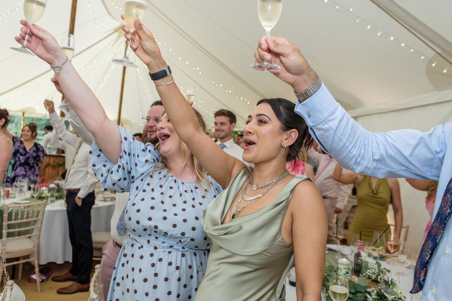 Guests raising champagne glasses in a toast at an elegant wedding reception under a marquee tent.