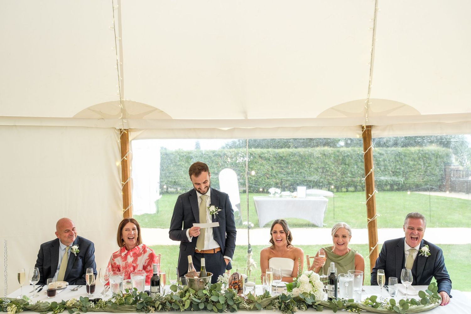 Wedding party seated at head table in a marquee, with best man standing giving a speech during the reception.
